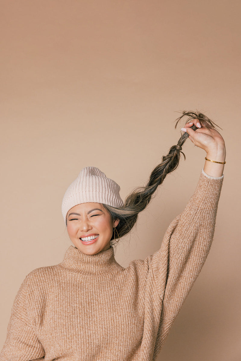 Smiling woman holding up a braided ponytail while wearing a beige knit sweater and light pink beanie against a beige background