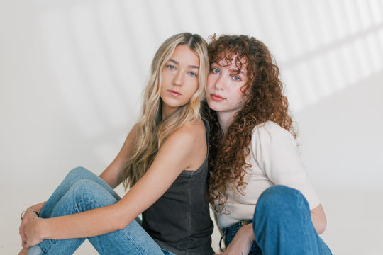 Two women sitting close together, one with long straight blonde hair and one with curly auburn hair, posing against a bright studio background