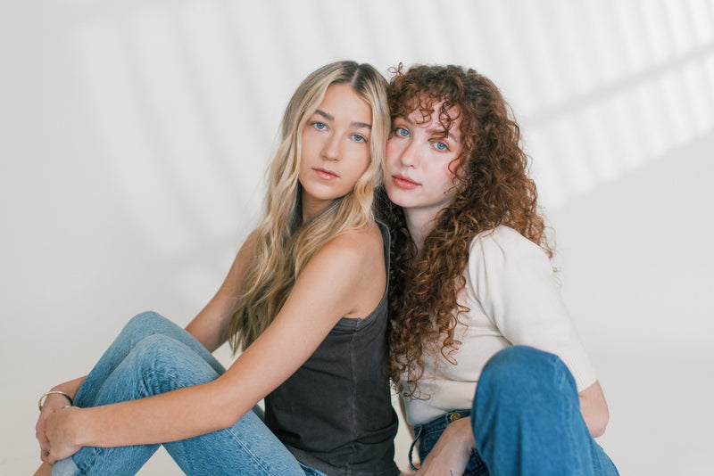 Two women sitting close together, one with long straight blonde hair and one with curly auburn hair, posing against a bright studio background