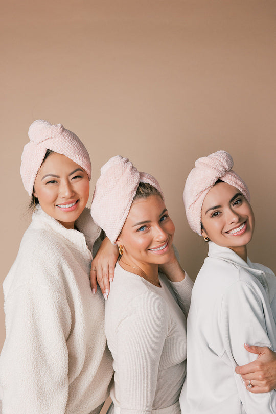 Three women wearing pink towel turbans, smiling and posing together against a beige background