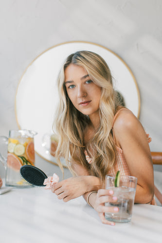 Young woman with long blonde hair holding a glass of water and a hairbrush, sitting at a table with a pitcher of lemon water, reflecting hair care and hydration theme.