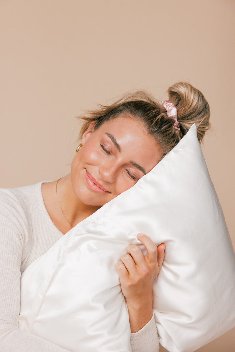 A woman with a pink scrunchie hugging a white pillow with eyes closed, smiling peacefully as if napping or resting.
