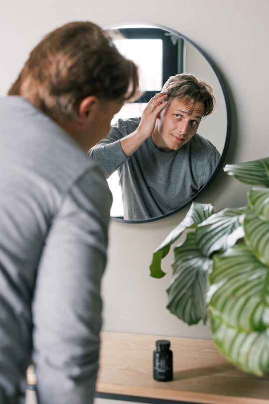 Man examining his hair in a round mirror, concerned about hair loss, with a bottle of hair supplement on the table nearby.