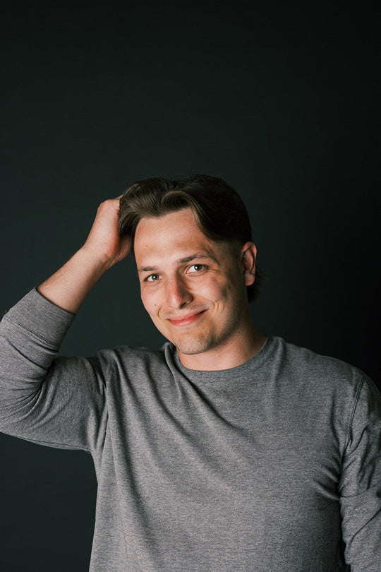 Smiling man with medium-length brown hair touching his head against a dark background