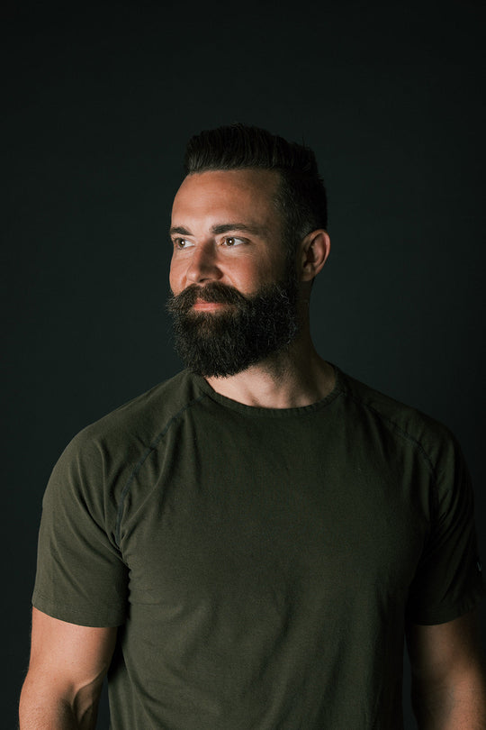 Portrait of a man with styled dark hair and a full beard wearing a dark green t-shirt against a dark background, illustrating male hair care.