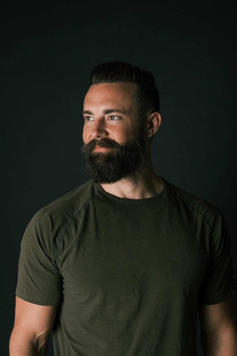 Man with styled dark hair and full beard looking to the side against a dark background