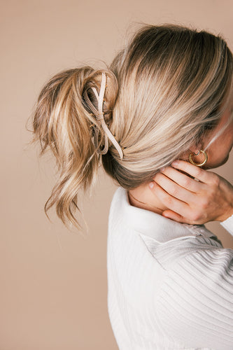 Woman with blonde hair styled in a loose bun secured with a beige hair clip, wearing a white top and gold hoop earrings, touching her neck.