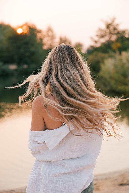 Woman with long, flowing blonde hair standing by a lake at sunset, wearing a white off-shoulder top.