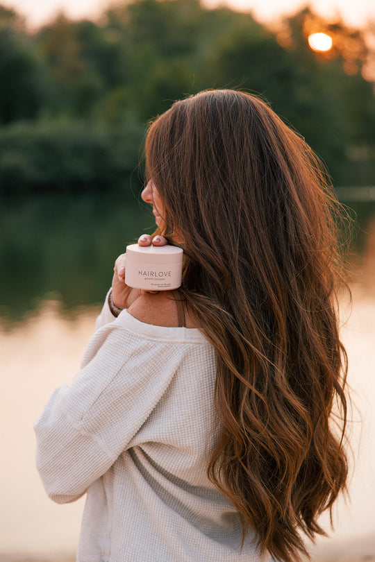 Woman with long brown hair holding a container labeled 'HAIRLOVE growth complex' near her shoulder, outdoors by a body of water at sunset.
