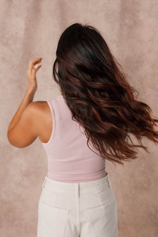 Person with long, dark wavy hair wearing a light pink sleeveless top and white pants, shown from behind against a neutral background.