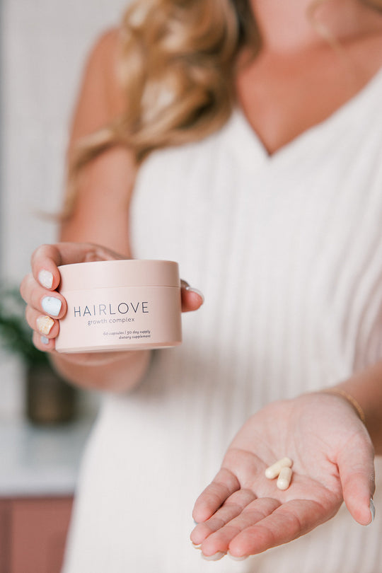 Person holding a container labeled 'HAIRLOVE growth complex' and showing three capsules in their open palm, wearing a white sleeveless top.