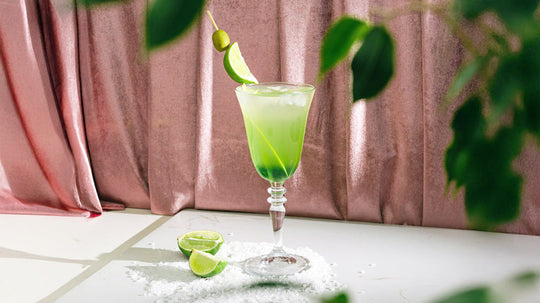 Green margarita in a stemmed glass garnished with a lime wedge and two green olives on a skewer, with lime wedges and salt scattered on the white surface, pink curtain background.