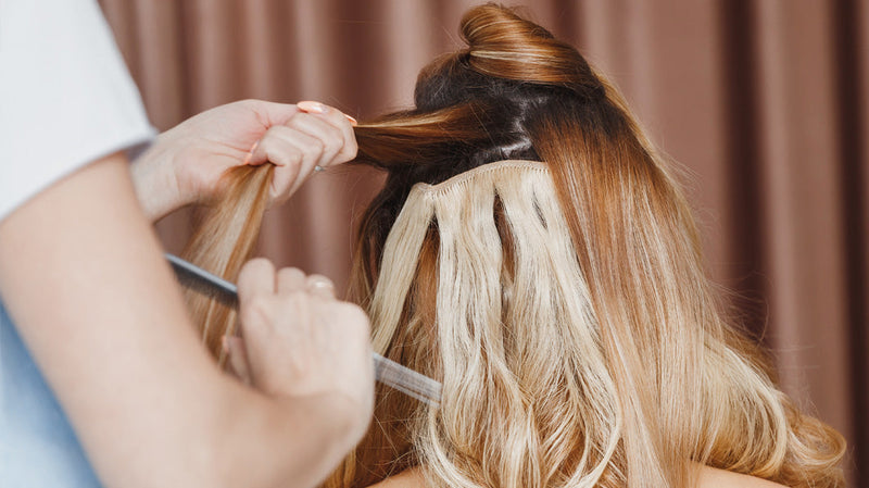 Hairstylist applying tape-in hair extensions to the back of a client’s blonde hair in a salon