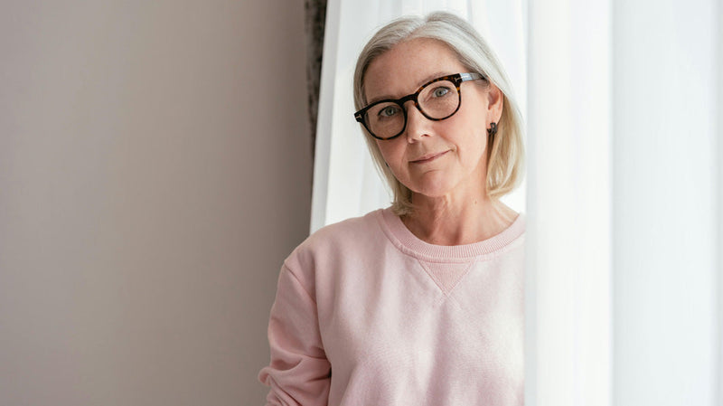 Middle-aged woman with gray hair and glasses standing by a window in a pink sweater, looking thoughtful