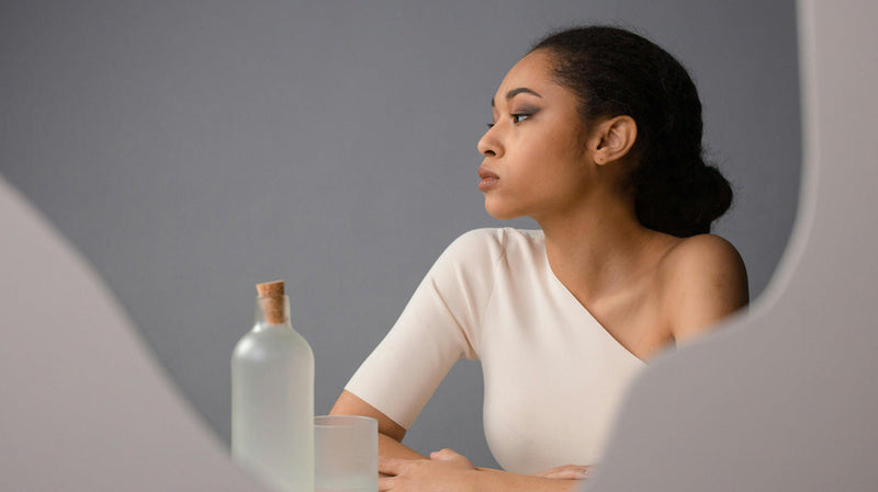 Profile of a woman with dark hair pulled back, looking thoughtfully to the side, seated at a table with a glass bottle and glass in front of her, against a gray background.
