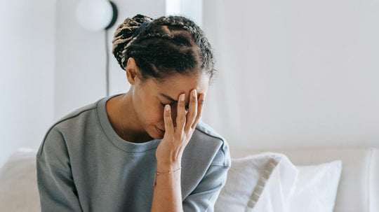 Woman with braided hair sitting on a bed, holding her forehead and looking distressed in a bright bedroom