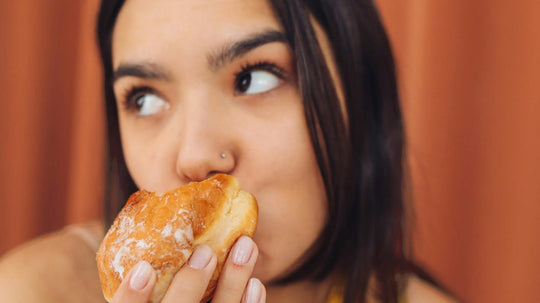 Person bringing a glazed donut to their mouth, looking thoughtful against a warm brown curtain background