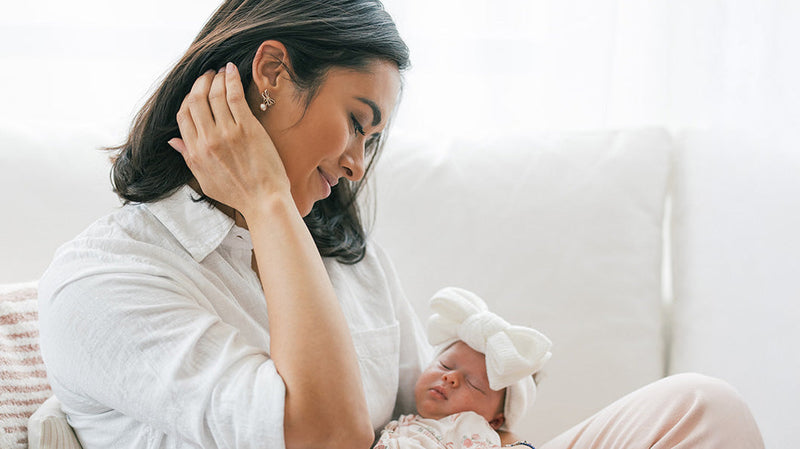 Mother gently holding her sleeping newborn baby wrapped in a blanket with a large bow headband, sitting on a couch in soft natural light.