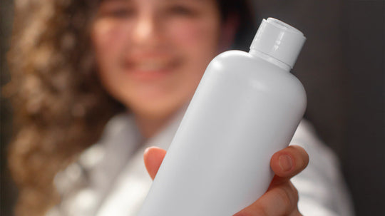 Person holding a white shampoo bottle toward the camera, smiling with a blurred background