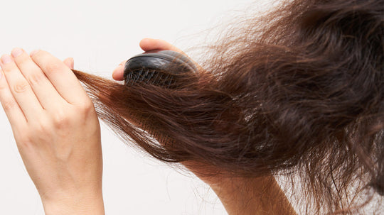 Person brushing frizzy brown hair with a black paddle brush against a light background