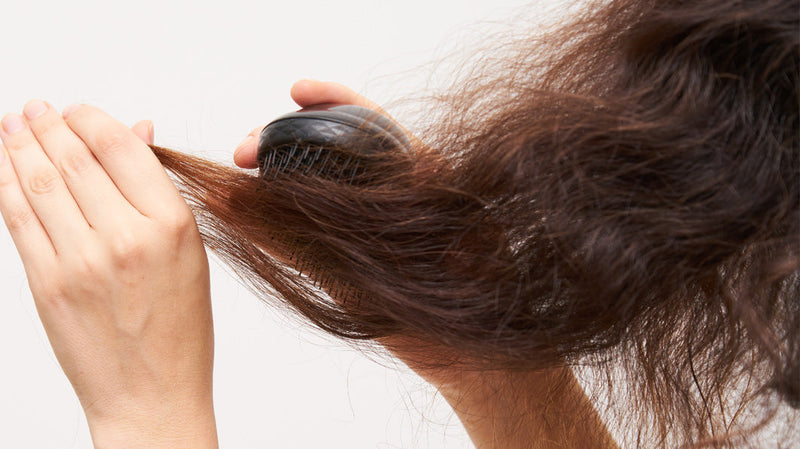 Person brushing frizzy brown hair with a black paddle brush against a light background