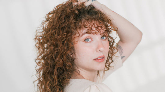 Woman with curly auburn hair touching her hair and looking over her shoulder against a light background