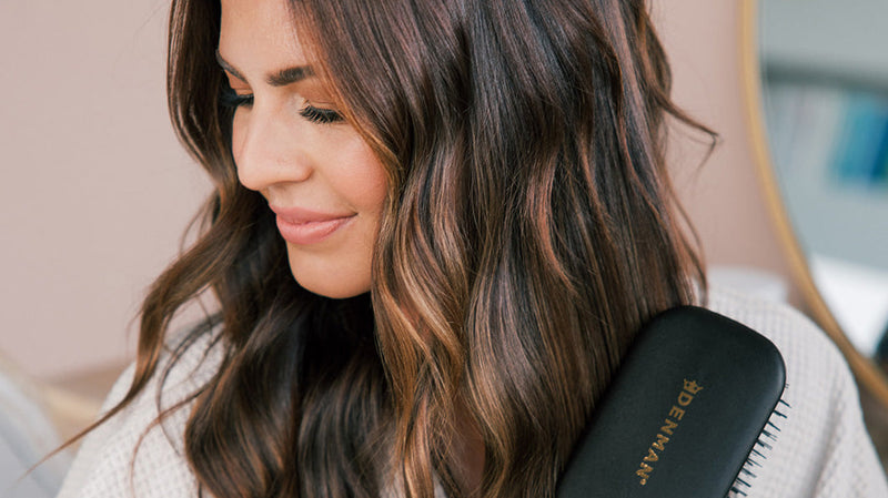 Woman with long brown hair holding a Denman hairbrush, smiling and looking down, illustrating hair care for volume and revival.