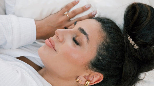 Woman with dark hair tied back sleeping peacefully on a white pillow, wearing a white robe and gold hoop earrings