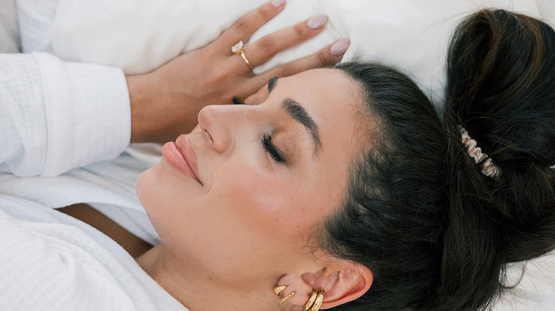 Woman with dark hair tied back sleeping peacefully on a white pillow, wearing a white robe and gold hoop earrings