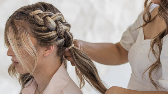 Close-up of a hairstylist braiding a blonde woman’s highlighted hair into a loose side braid in a neutral studio setting