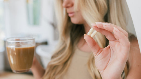 Blonde woman holding a glass cup of coffee in one hand and two supplement capsules in the other, softly lit indoor kitchen background