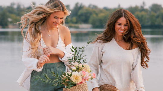 Two women strolling along a lake at golden hour, smiling and showing summer hairstyles, one holding a woven basket of flowers