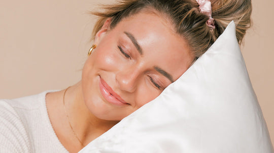 Woman with closed eyes resting her head on a white pillow, smiling gently, with hair tied back by a pink scrunchie.