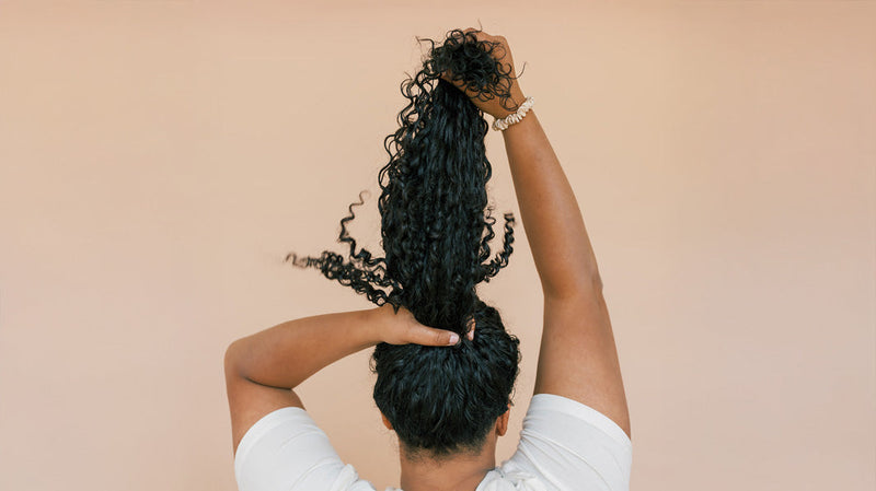 Woman lifting long curly black hair into a high ponytail, viewed from behind against a beige background
