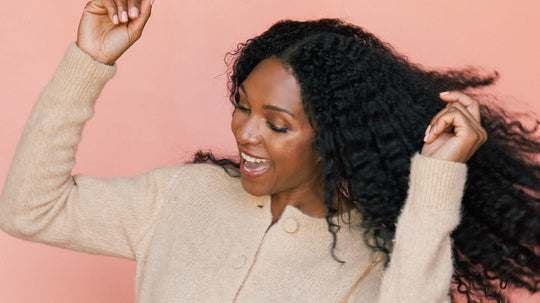 Woman with long curly hair dancing and smiling against a pink background, wearing a beige sweater