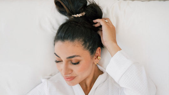 Woman with dark hair resting on a white pillow, eyes closed and hand near her head in a bright bed setting