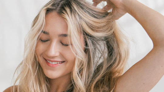 Smiling woman with long blonde highlighted hair touching her hair with eyes closed against a light neutral background