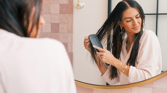 Woman in a pink robe brushing wet hair with a paddle brush while looking into a bathroom mirror