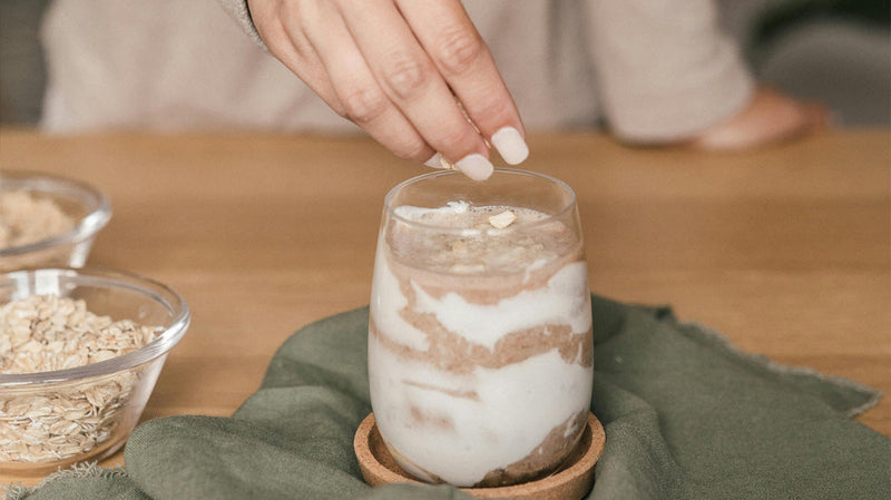 Hand sprinkling oats onto a layered yogurt-and-cocoa parfait in a glass, bowls of oats beside it on a green cloth