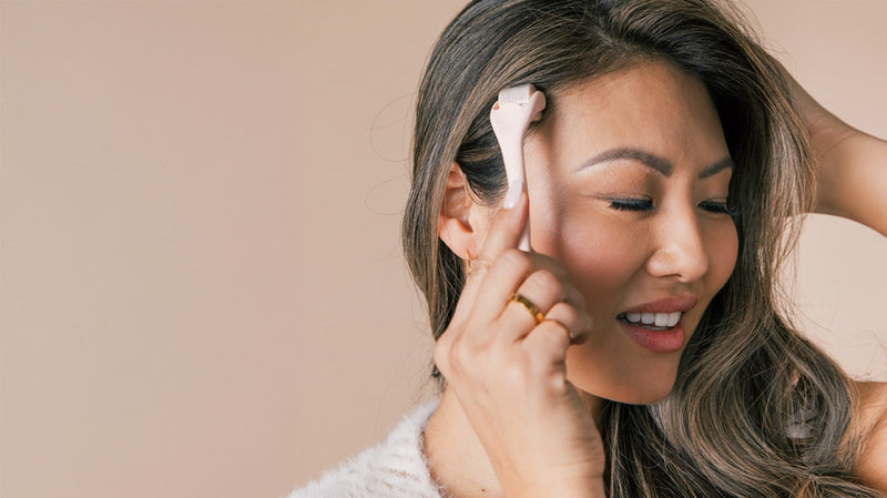 Woman using a pink handheld facial massager near her ear, smiling against a beige background