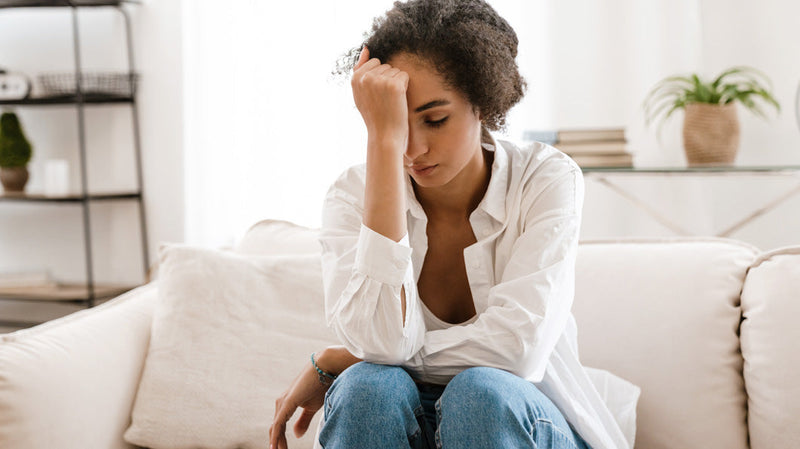 Seated woman on a couch, looking distressed and holding her forehead, conveying worry about hair loss.