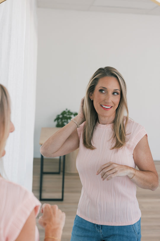Woman smiling at her reflection while smoothing shoulder-length blond hair, wearing a pink pleated top and jeans