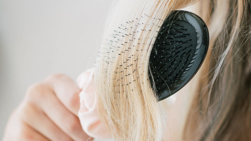 Blonde hair tangled in a black paddle brush as a hand holds a pale pink cloth