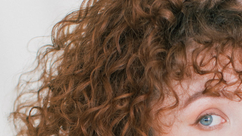 Close-up of curly reddish-brown hair with textured ringlets and a partial blue eye at right.