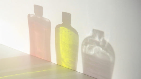 Three translucent shampoo bottles in a row showing pink, yellow, and clear liquids and casting colored shadows on a white wall