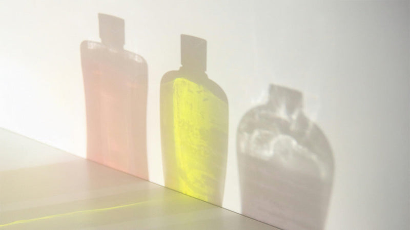 Three translucent shampoo bottles in a row showing pink, yellow, and clear liquids and casting colored shadows on a white wall