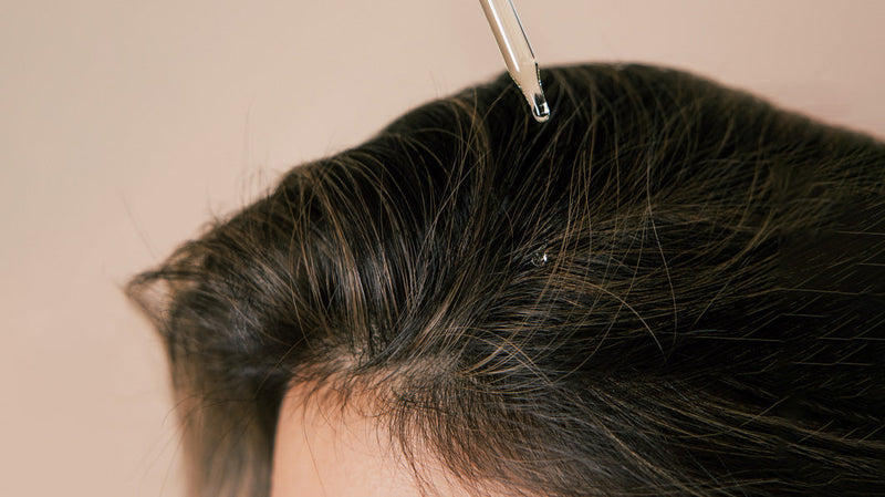 Close-up of a scalp and hair part with a glass dropper releasing a droplet