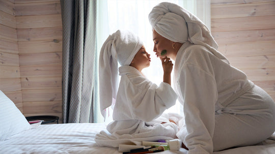 Mother and child in bathrobes and towel turbans playfully applying makeup on a bed, brushes and cosmetics in foreground.