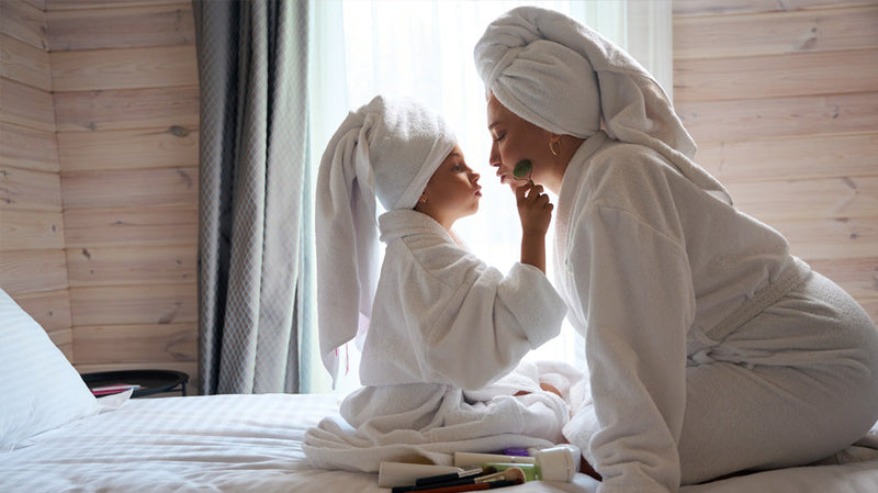 Mother and daughter playfully applying makeup on a bed while wearing bathrobes and towel turbans