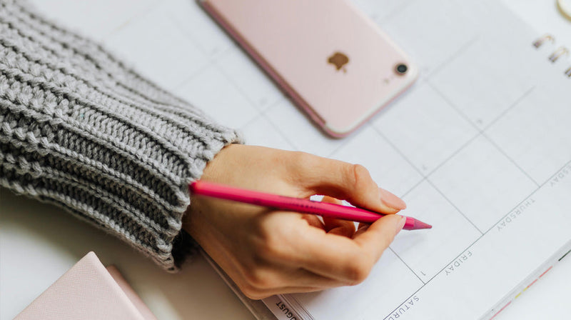 Hand writing in a weekly planner with boxes labeled FRIDAY SATURDAY, pink pencil, iPhone and grey sweater sleeve.